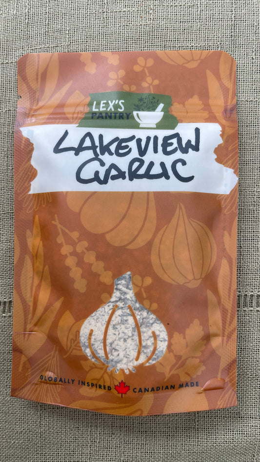 A stand-up pouch of Lex's Pantry Lakeview Garlic Herb & Spice Blend on a textured beige fabric background. The packaging is kraft-brown with a clear garlic bulb window showing the mixed seasoning inside and a white label across the middle.