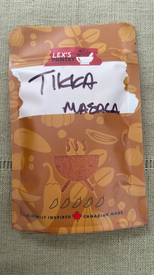 A stand-up pouch of Lex's Pantry Tikka Masala , featuring a brown design with illustrations of spices and a white label area with handwritten text, placed on a textured beige fabric background.