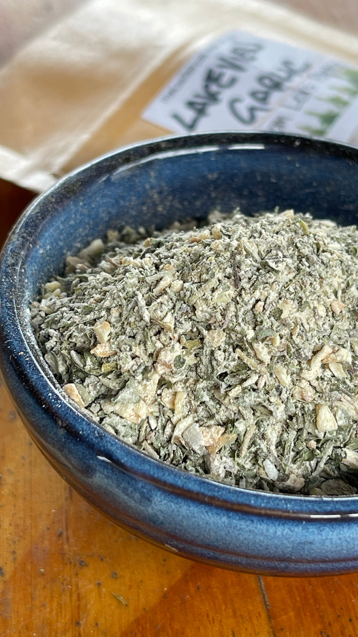 A closeup of Lakeview Garlic & herb blend by Lex's Pantry in a small blue bowl on a wooden surface with a label in the background