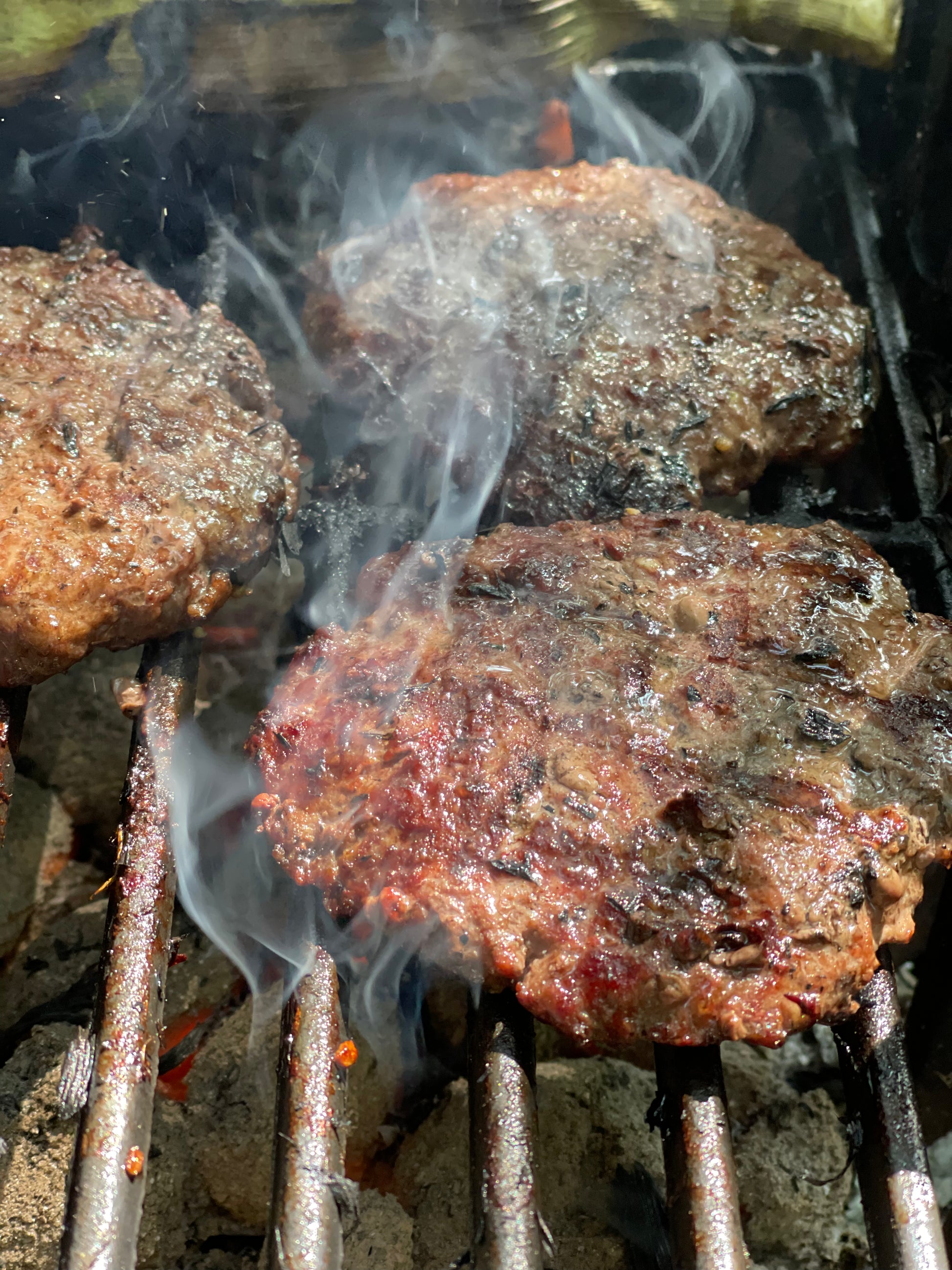 burgers made with Bay Burger Blend From Lex's Pantry on the grill over red hot coals with smoke tendrils flowing up