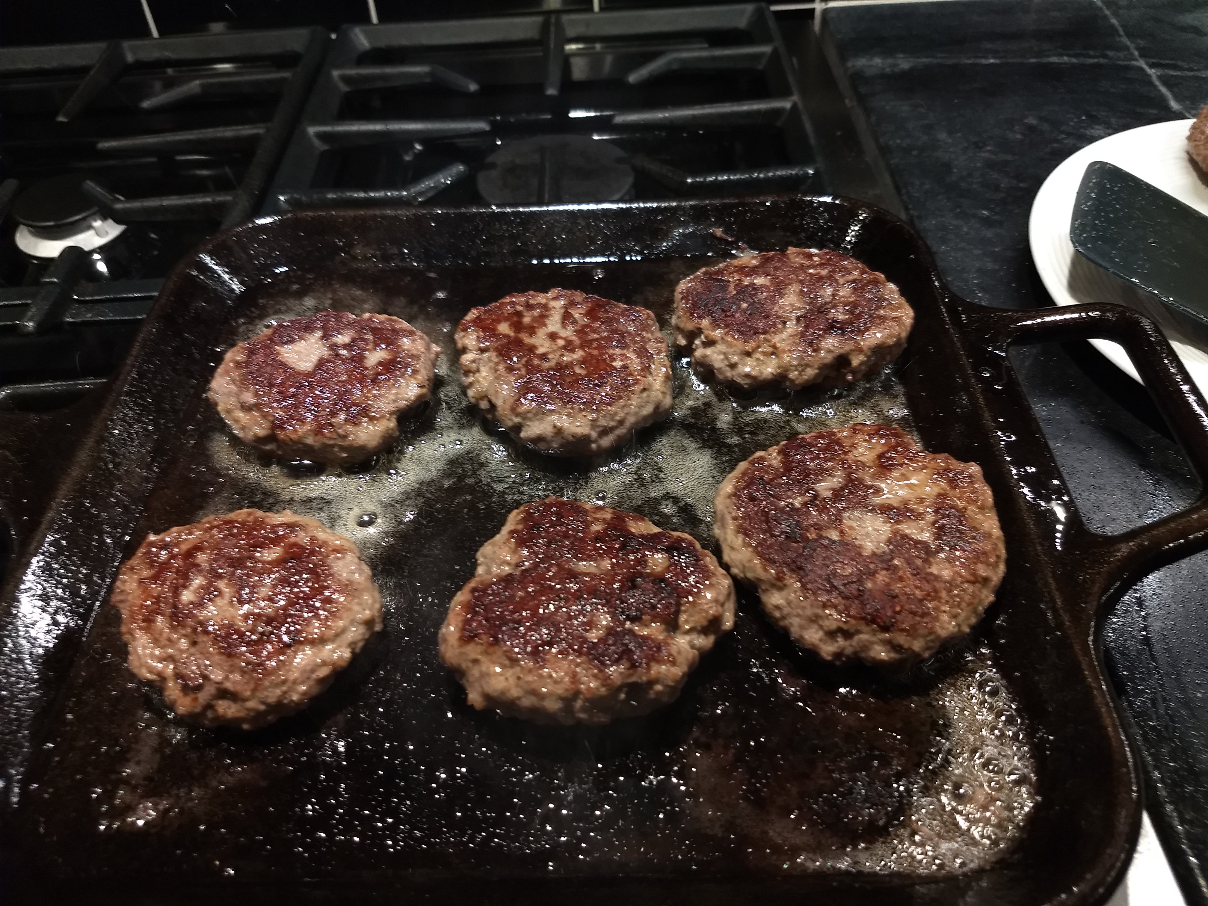 6 burger patties on a square cast iron griddle on a gas stove top