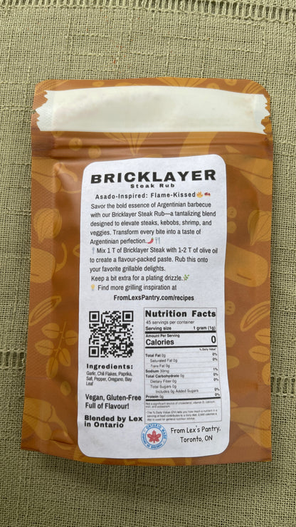 The back of a bag of Lex's Pantry Bricklayer Steak Rub against a textured olive green fabric background. The packaging is kraft brown with a large white label showing product details, a QR code, and nutrition facts and a Made in Ontario label.