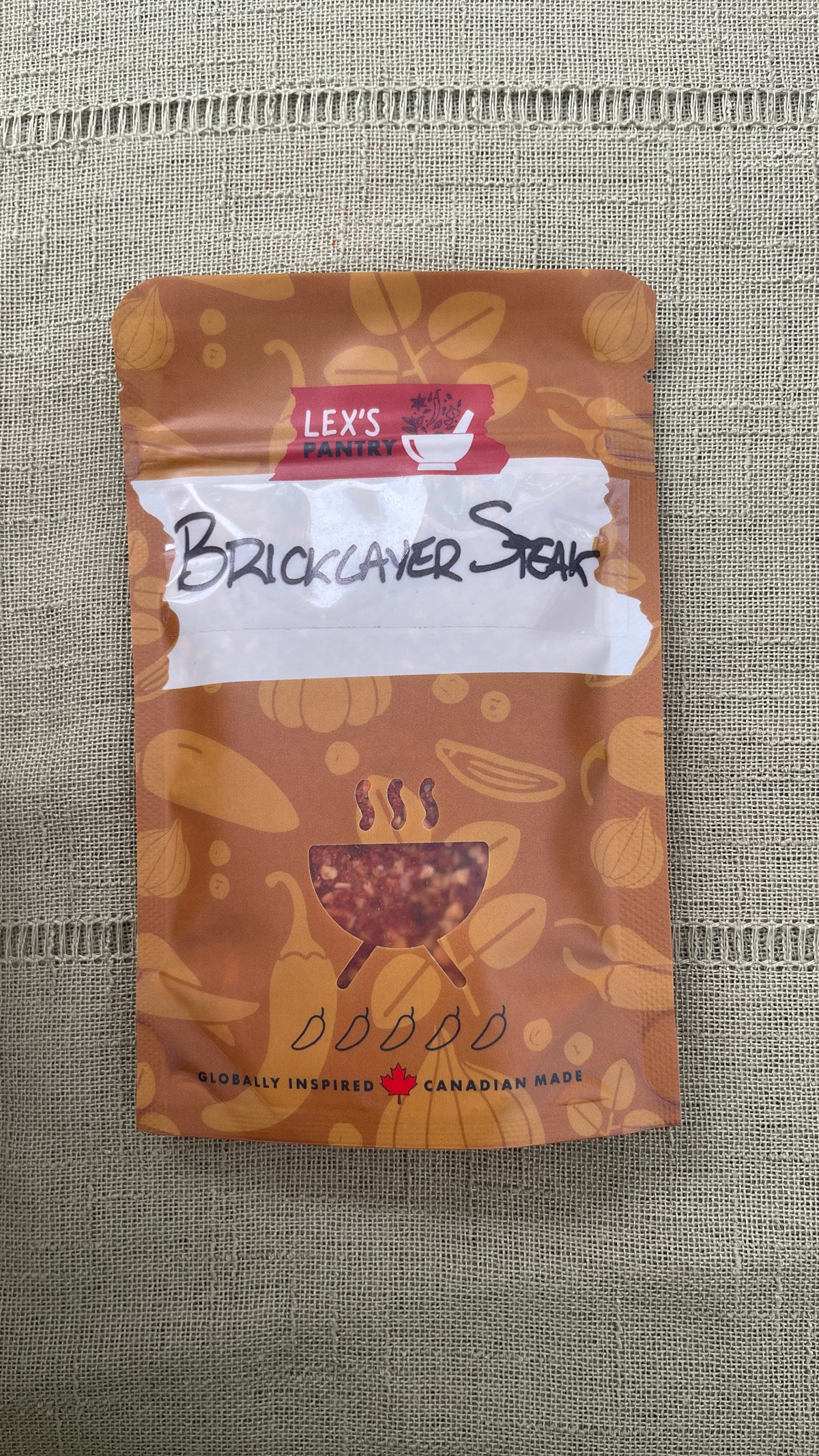 A stand-up pouch of Lex's Pantry Smokehouse Bricklayer Steak Rub, featuring an kraft-brown patterned design with illustrations of spices and herbs and a BBQ cutout graphic.