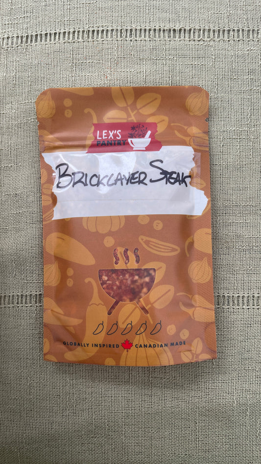 A stand-up pouch of Lex's Pantry Smokehouse Bricklayer Steak Rub, featuring an kraft-brown patterned design with illustrations of spices and herbs and a BBQ cutout graphic.