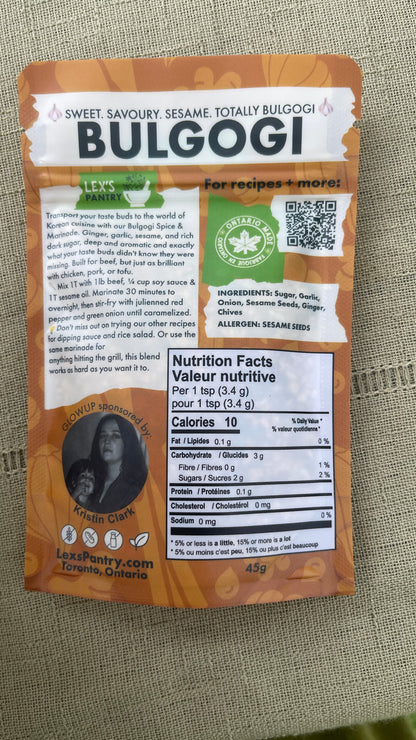 The back of a bag of Lex' Pantry Bulgogi seasoning & marinade mix on a textured fabric background. The packaging is kraft-brown with white and black text, featuring a circular photo of Lex & her mom, a QR code, and a prominent white nutrition facts label.