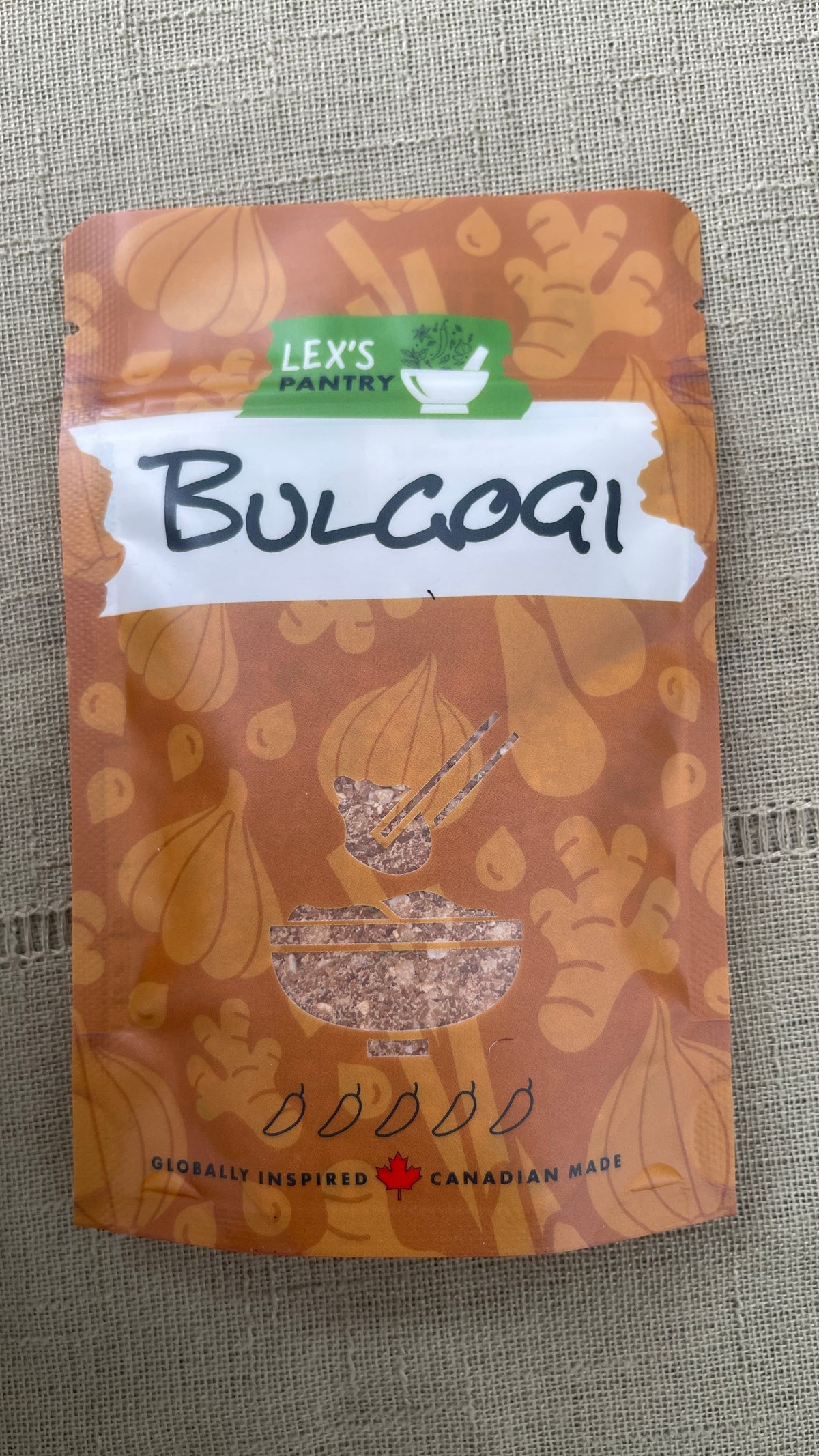 A stand-up pouch of Lex's Pantry Bulgogi seasoning & marinade, featuring an kraft-brown patterned background with illustrations of garlic cloves, ginger, and sesame seeds, and a white label with black handwritten text, placed on a textured  kraft-brown surface.
