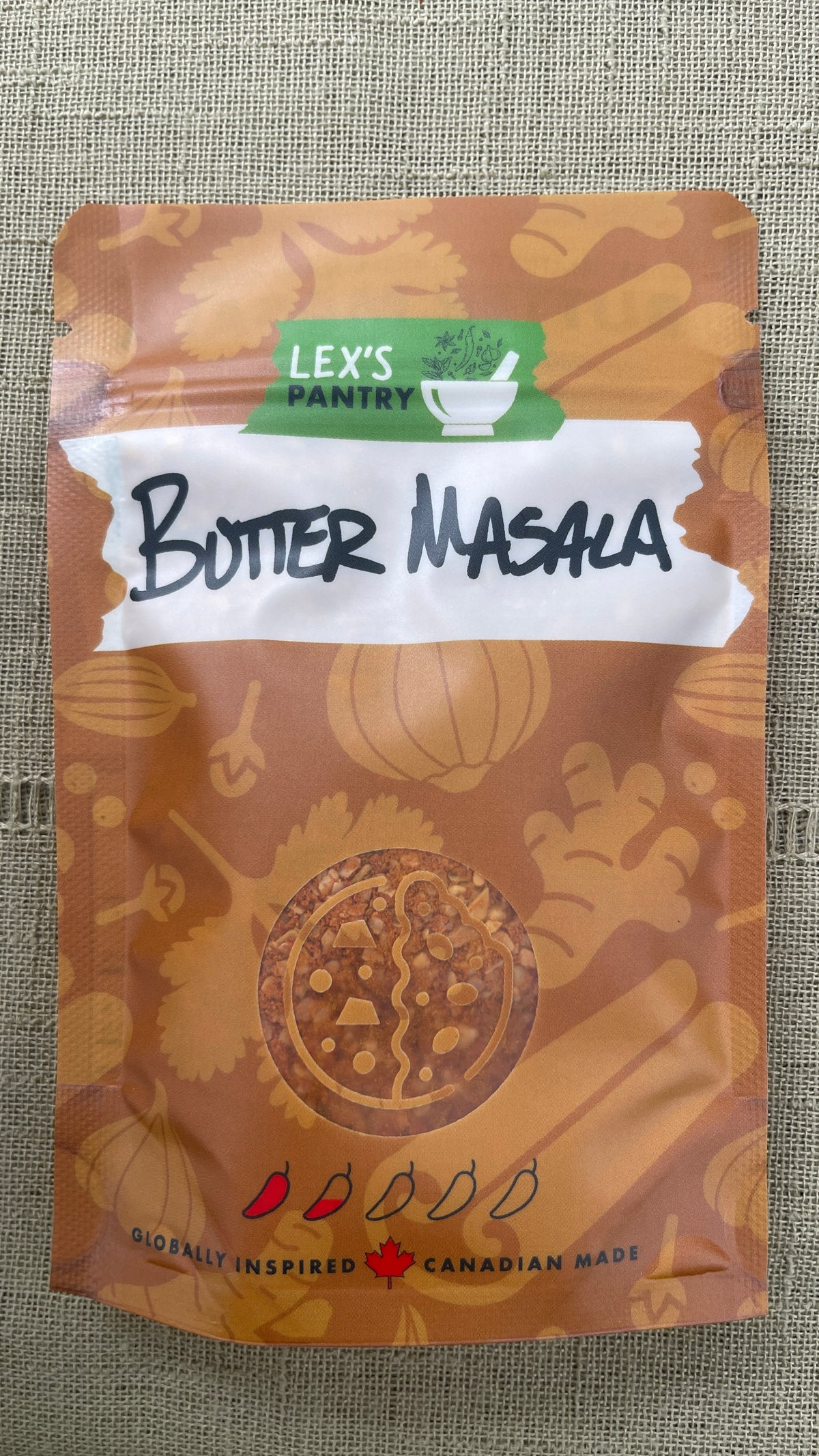 A stand-up pouch of Lex's Pantry Butter Masala spice mix, featuring a warm brown and orange patterned design with a white label and green brand logo, set against a textured beige fabric background.