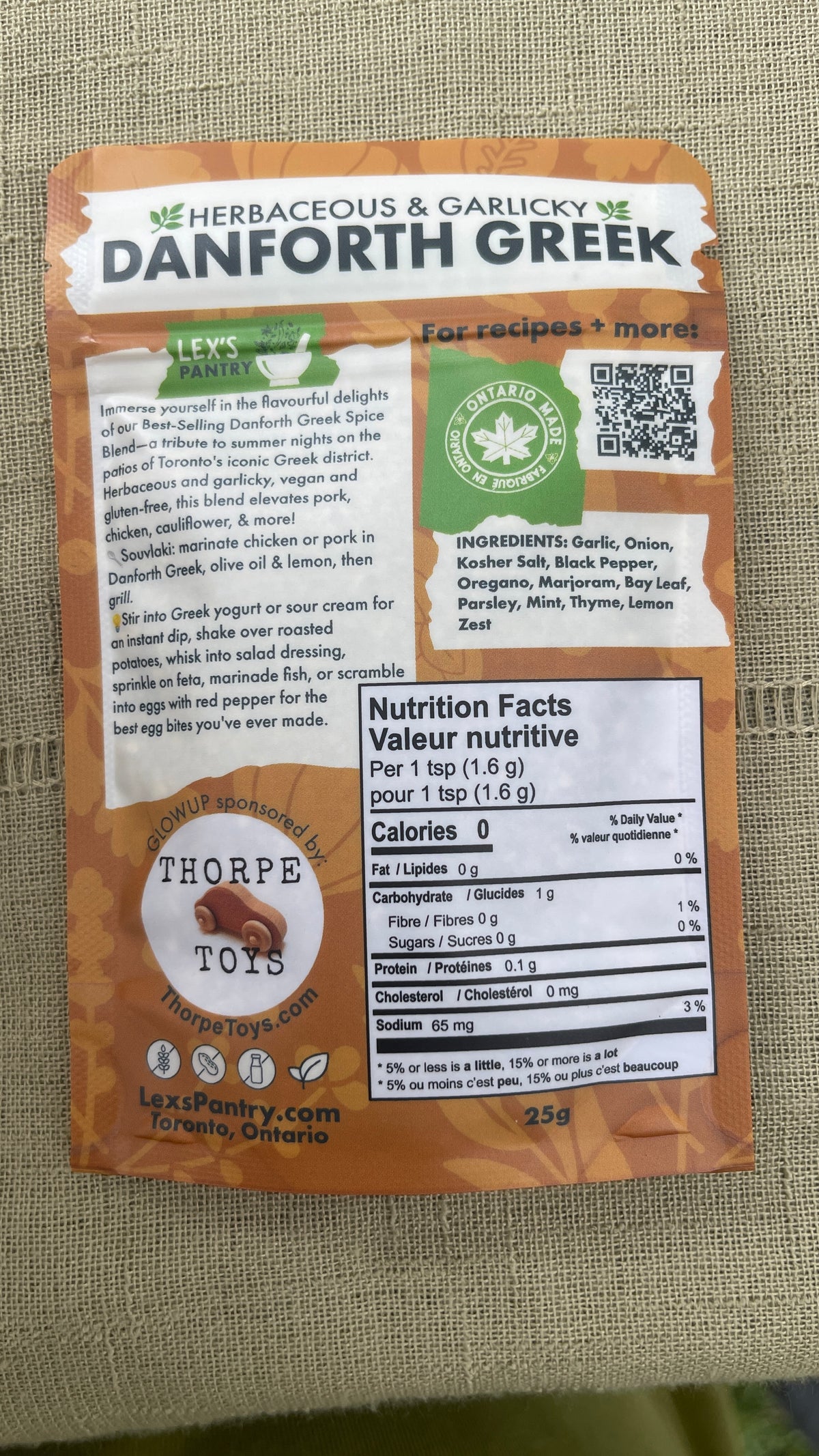 A bag of Lex's Pantry  Danforth Greek described as herbaceous & Garlicky. The packaging is kraft brown with white and green accents. Featuring nutrition details, description, ingredients, a QR code to recipes, an Ontario Made logo and Lex's Pantry's Glowup sponsor - 'Thorpe Toys' brand logo on a textured beige fabric background.