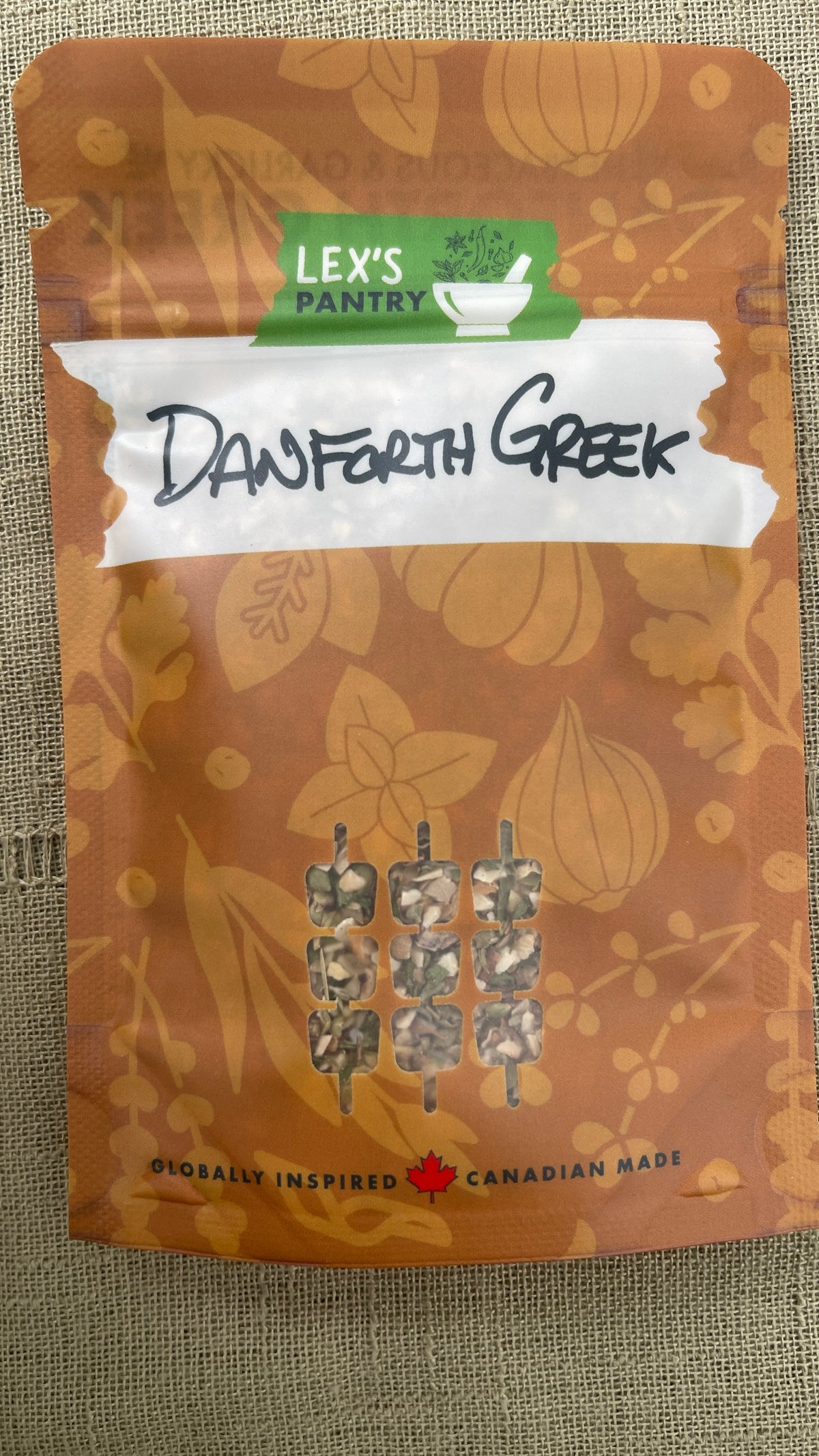 A standup bag of Lex's Pantry Danforth Greek seasoning and marinade. A brown kraft-style pouch with a white tape graphic with the flavour handwritten across it, and small skewered food cutout on the front, photographed against a textured fabric background.