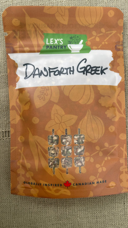 A standup bag of Lex's Pantry Danforth Greek seasoning and marinade. A brown kraft-style pouch with a white tape graphic with the flavour handwritten across it, and small skewered food cutout on the front, photographed against a textured fabric background.