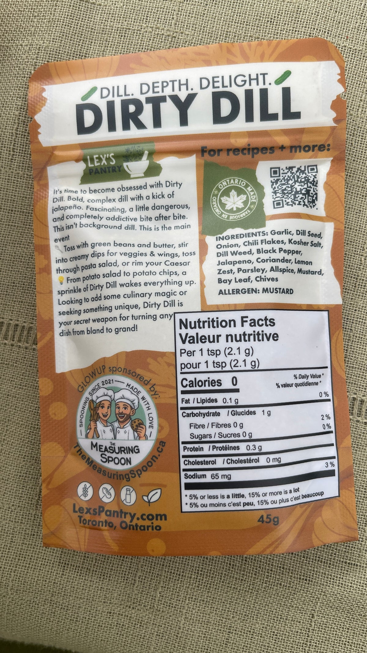 The back of a 45g "regular" bag of Lex's Pantry Dirty Dill spice blend, featuring a kraft brown design with detailed product information, ingredients, and a QR code on a textured fabric background.