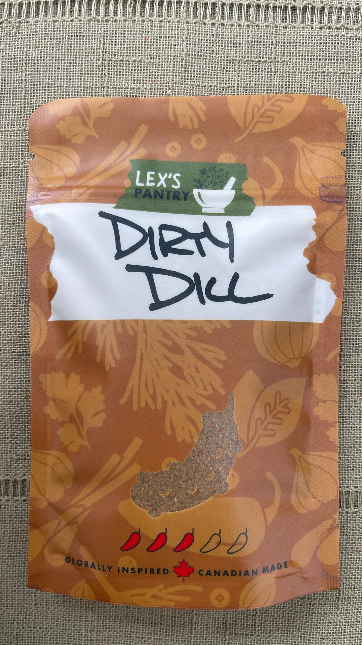 A stand-up pouch of Lex's Pantry 'Dirty Dill' seasoning, featuring a brown and orange botanical pattern with a white label and black handwritten text, set against a textured beige burlap background.