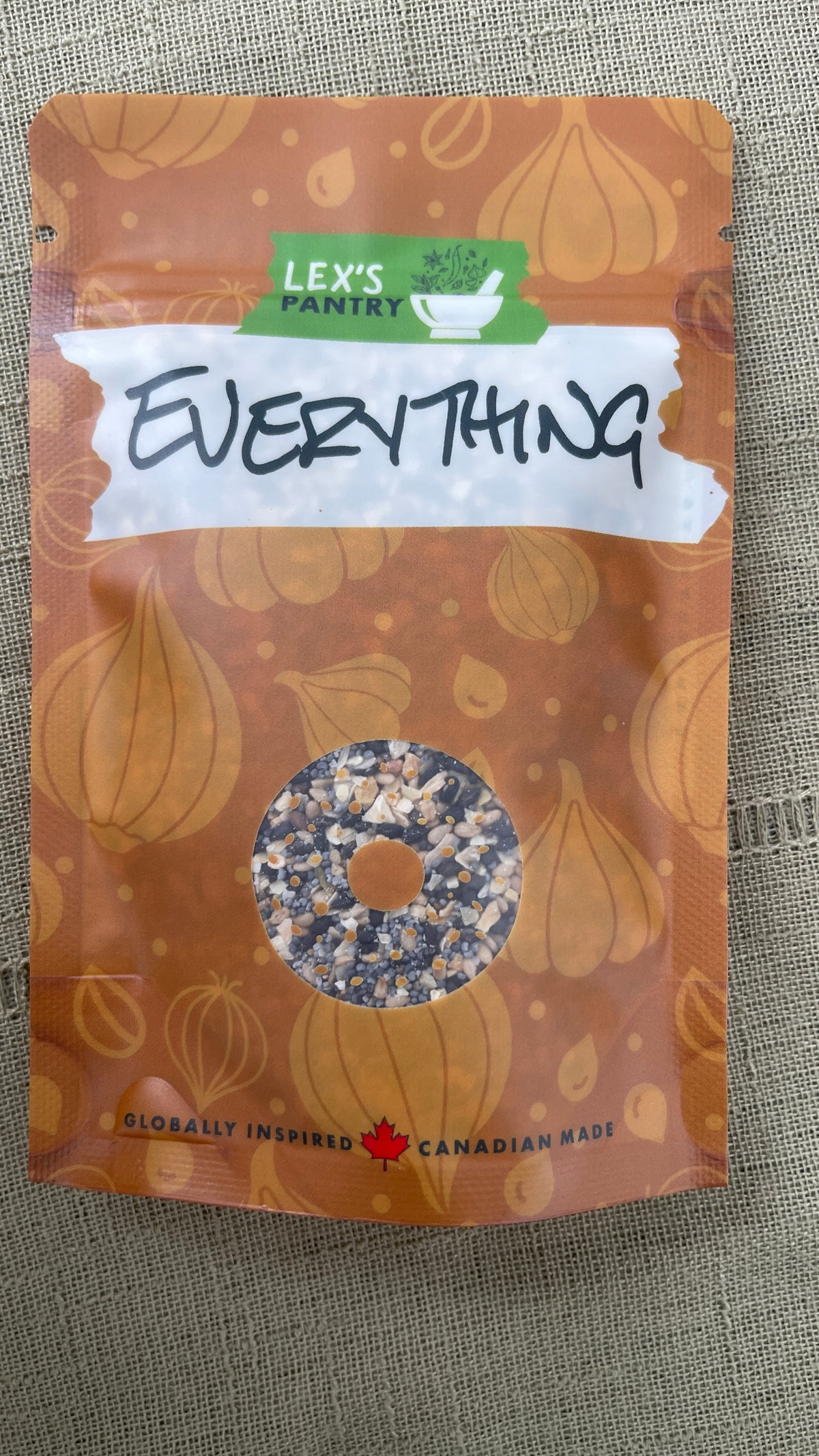 A stand-up pouch of Lex's Pantry Everything Bagel seasoning mix, featuring an kraft-brown pattern of garlic, sesame seeds, poppy seeds and onion and a clear bagel shaped window showing the mixed seasoning inside. The brand logo is green and white, and the packaging is photographed on a textured beige fabric background.