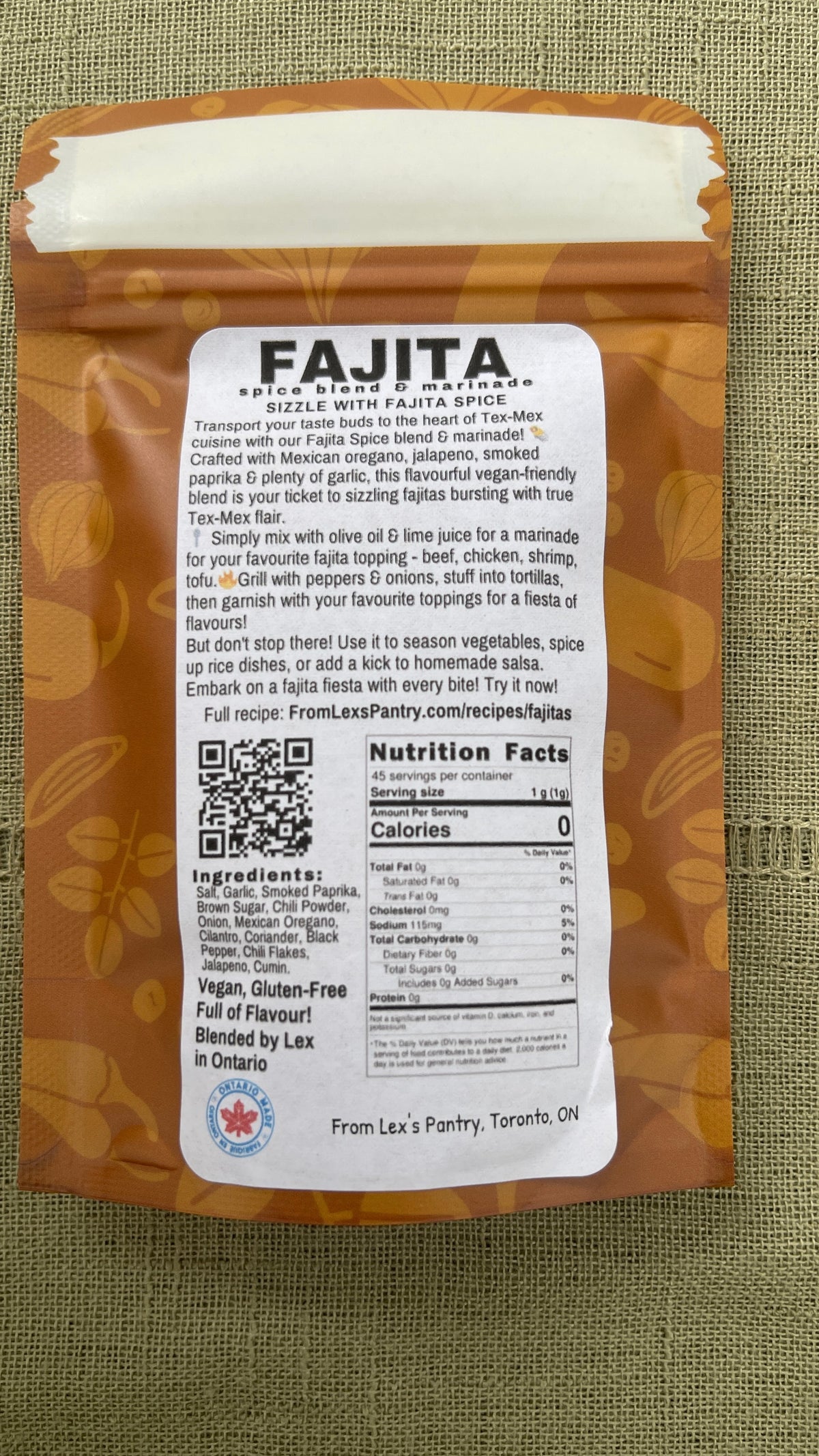 The back of a bag of Lex's Pantry's Fajita Spice Blend on a textured green fabric background. The package features a white label with product details, ingredients, and a QR code, set against a kraft-brown patterned background.