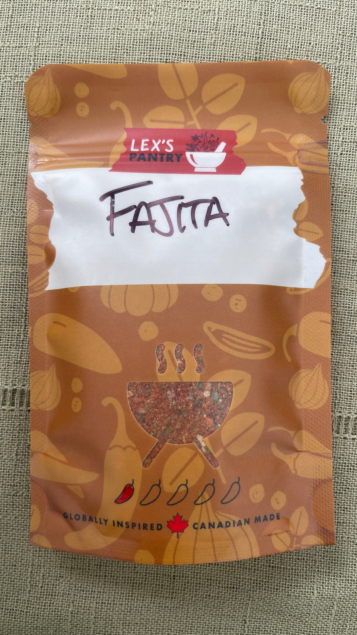 A stand-up pouch of Lex's Pantry Fajita seasoning mix, featuring an kraft-brown pattern of garlic, peppers, spices & herbs. A clear BBQ shaped window showing the mixed seasoning inside.  And a white label area with 'FAJITA' hand written in black.   The brand logo is red and white, and the packaging is photographed on a textured beige fabric background.