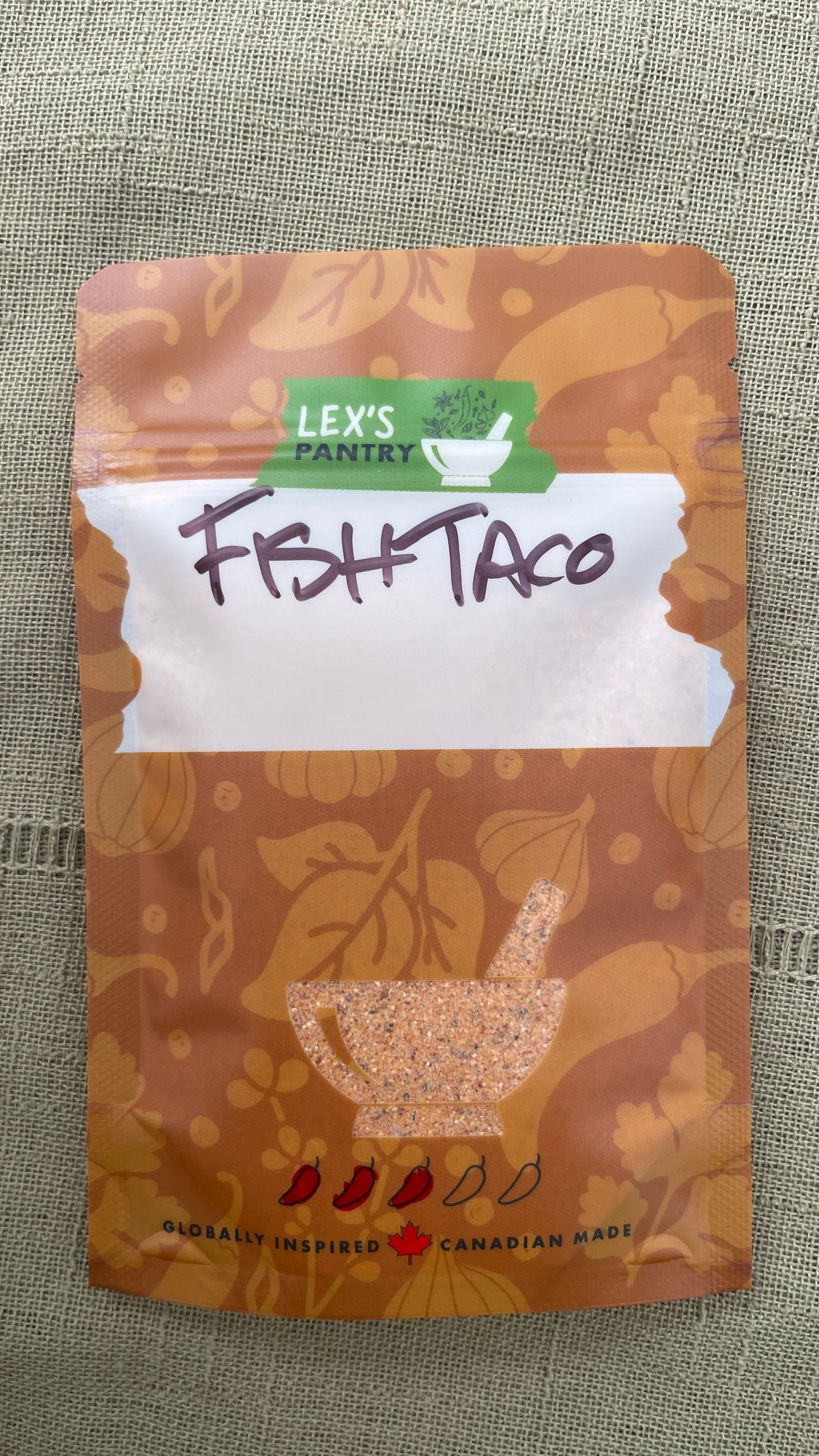 A stand-up pouch of Lex's Pantry Fish Taco seasoning and dredge mix, featuring a kraft brown patterned design  with illustrations of garlic, chiles and herbs, with a white label area and handwritten 'FISH TACO' text, and a clear mortar & pestle shaped window showing the mixed seasoning inside. The brand logo is green and white, and the packaging is photographed on a textured green fabric background
