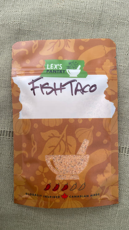 A stand-up pouch of Lex's Pantry Fish Taco seasoning and dredge mix, featuring a kraft brown patterned design  with illustrations of garlic, chiles and herbs, with a white label area and handwritten 'FISH TACO' text, and a clear mortar & pestle shaped window showing the mixed seasoning inside. The brand logo is green and white, and the packaging is photographed on a textured green fabric background