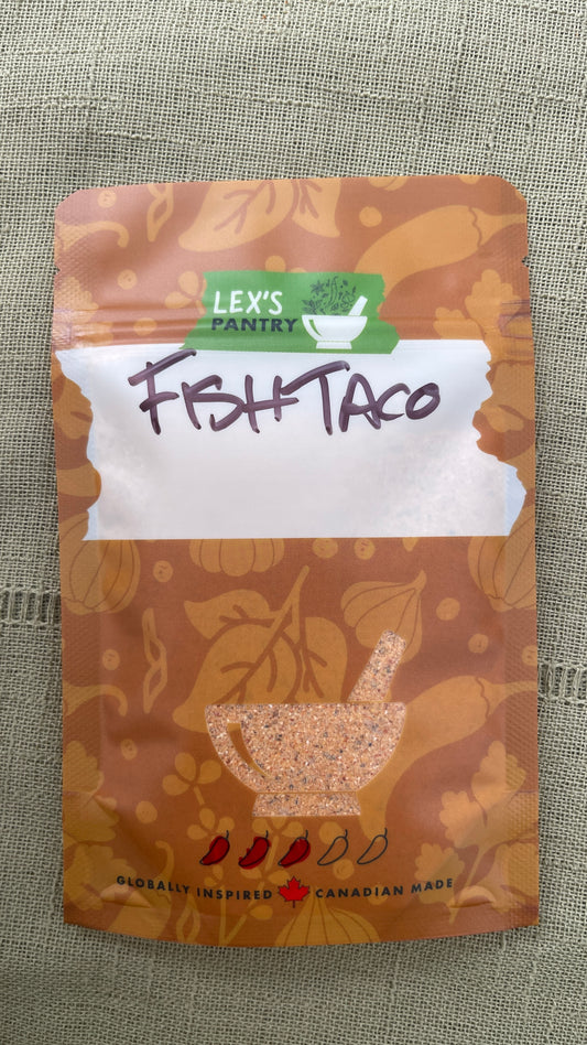 A stand-up pouch of Lex's Pantry Fish Taco seasoning and dredge mix, featuring a kraft brown patterned design  with illustrations of garlic, chiles and herbs, with a white label area and handwritten 'FISH TACO' text, and a clear mortar & pestle shaped window showing the mixed seasoning inside. The brand logo is green and white, and the packaging is photographed on a textured green fabric background