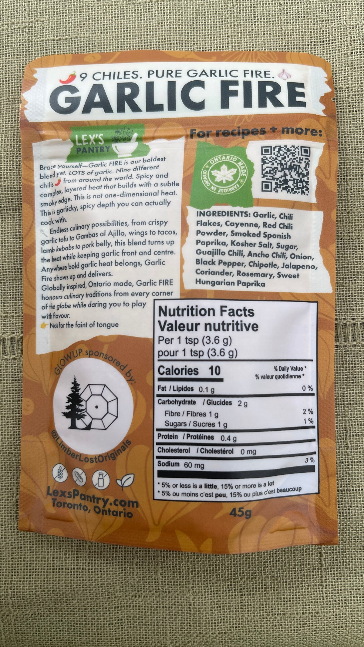 The back of a 45g regular bag of Lex's Pantry 'Garlic Fire' chili powder on a textured green fabric background. The packaging is kraft-brown with white and black text, featuring a prominent nutrition facts label, Ontario Made logo, and a QR code.