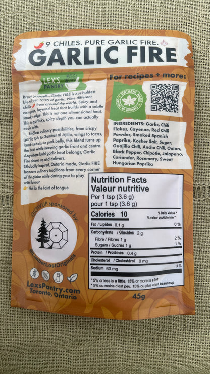 The back of a 45g regular bag of Lex's Pantry 'Garlic Fire' chili powder on a textured green fabric background. The packaging is kraft-brown with white and black text, featuring a prominent nutrition facts label, Ontario Made logo, and a QR code.