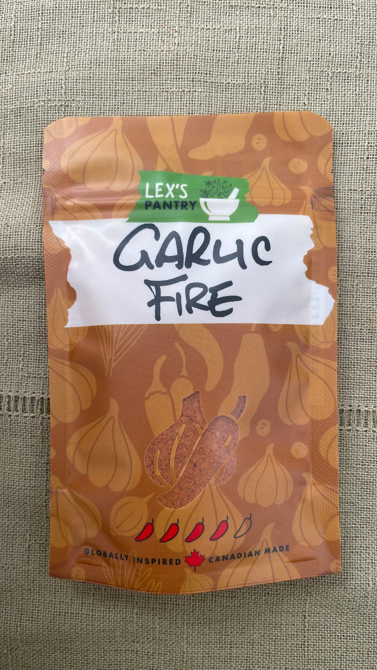 A stand-up pouch of Lex's Pantry Garlic Fire seasoning & spice mix, featuring a kraft brown garlic  and chili pattern.  A clear garlic & chili shaped window showing the mixed seasoning inside.  And a white label area with 'Garlic Fire' hand written in black. The brand logo is green and white, and the packaging is photographed on a textured green fabric background.
