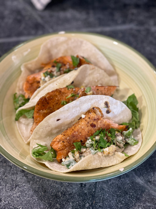 Three soft corn tacos filled with grilled fish made with Lex's Pantry Fish Taco spice & dredge, cilantro, corn salsa, and a creamy herb sauce, served on a light green plate against a dark gray stone surface.