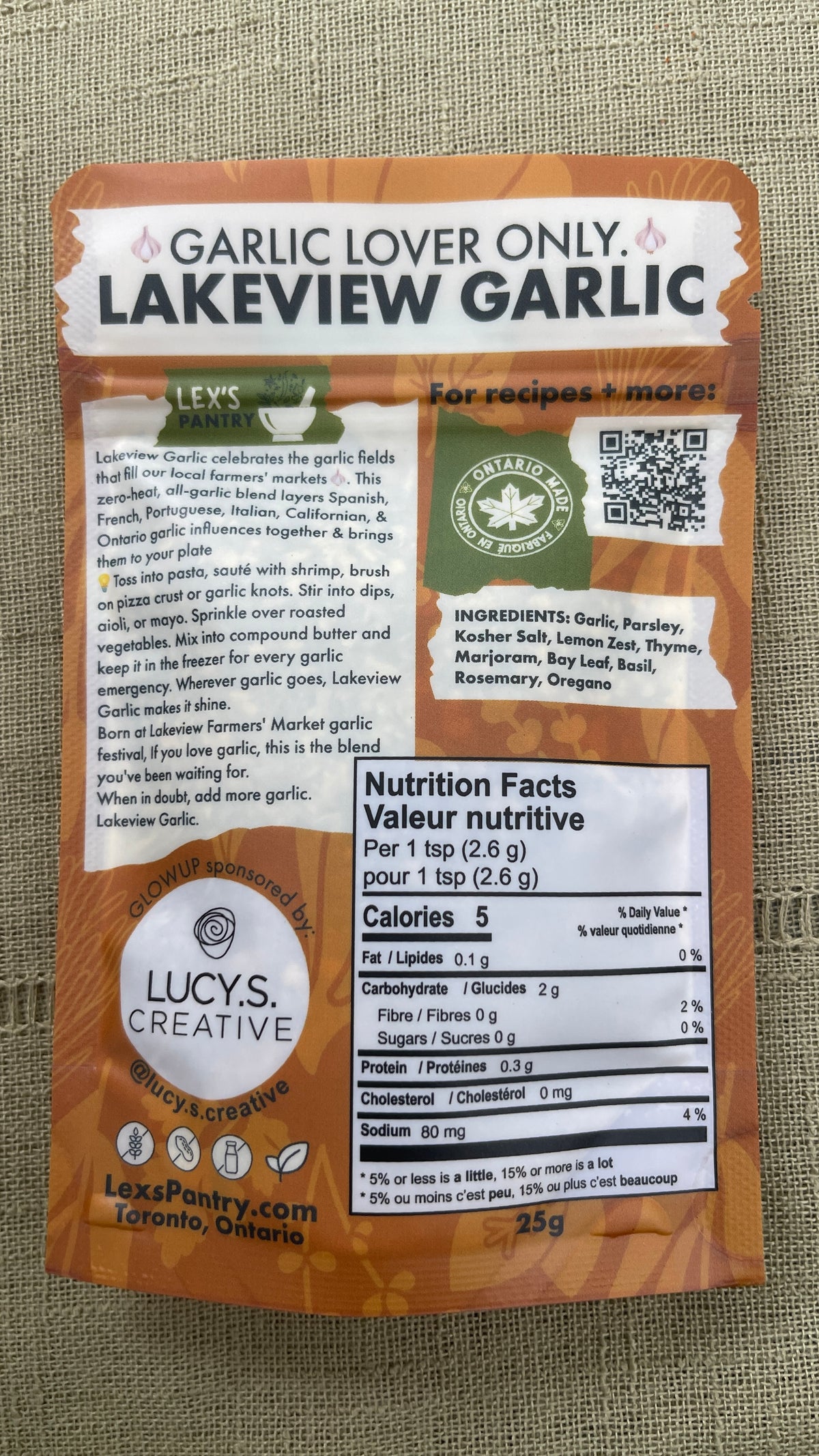 Back of the package photo of  Lakeview Garlic by Lex's Pantry, featuring a kraft brown label with product details, ingredients, and a nutrition facts panel on a textured beige fabric background.