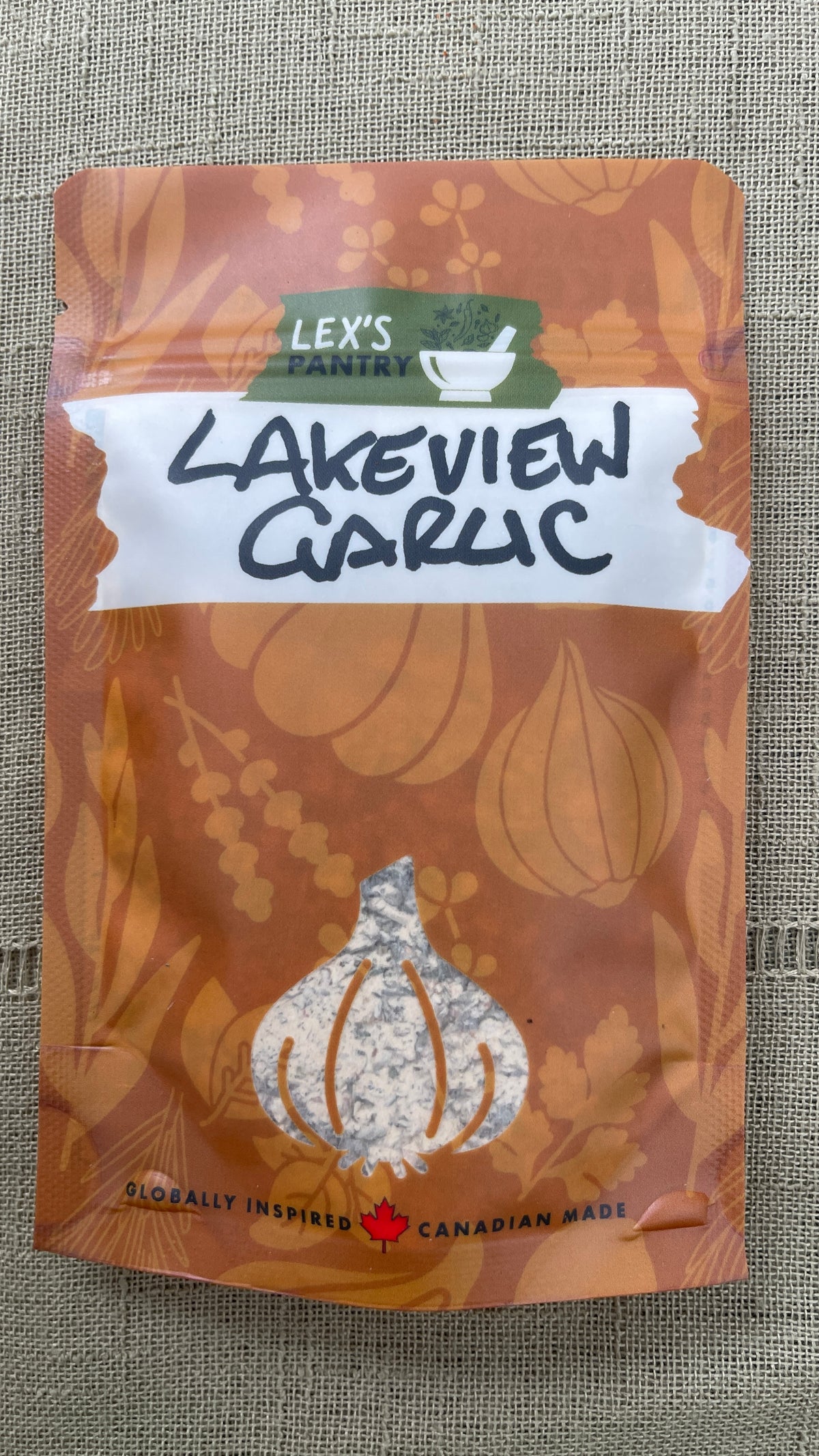 A stand-up pouch of Lex's Pantry Lakeview Garlic Herb & Spice Blend on a textured beige fabric background. The packaging is kraft-brown with a clear garlic bulb window showing the mixed seasoning inside and a white label across the middle.