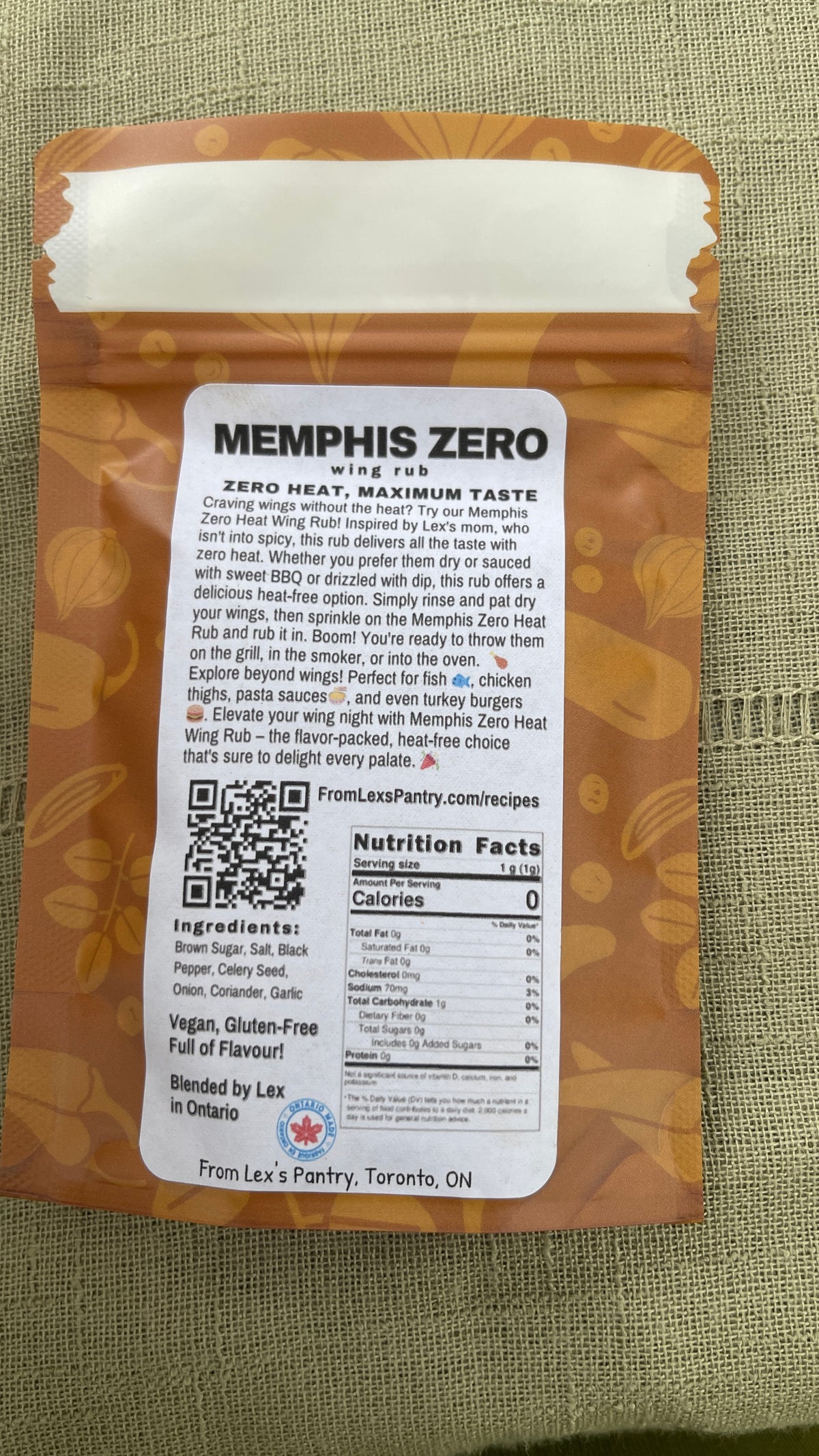 Back of a Memphis Zero Wing Rub bag from Lex's Pantry, showing the ingredients, nutrition facts, QR code, and brand information on a textured green fabric background.