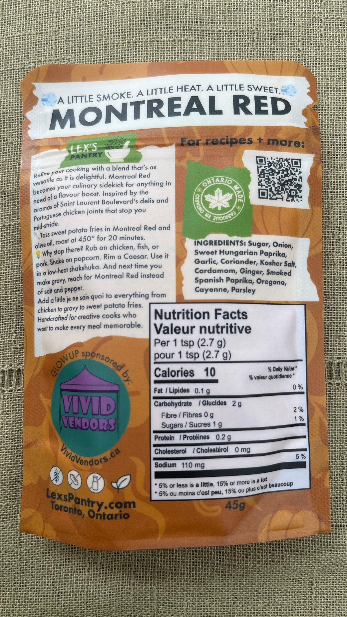 The back of a regular 45g bag of Lex's Pantry Montreal Red Chicken Rub featuring a warm kraft-brown background with a white nutrition facts label and a green brand logo, set against a textured beige fabric background.