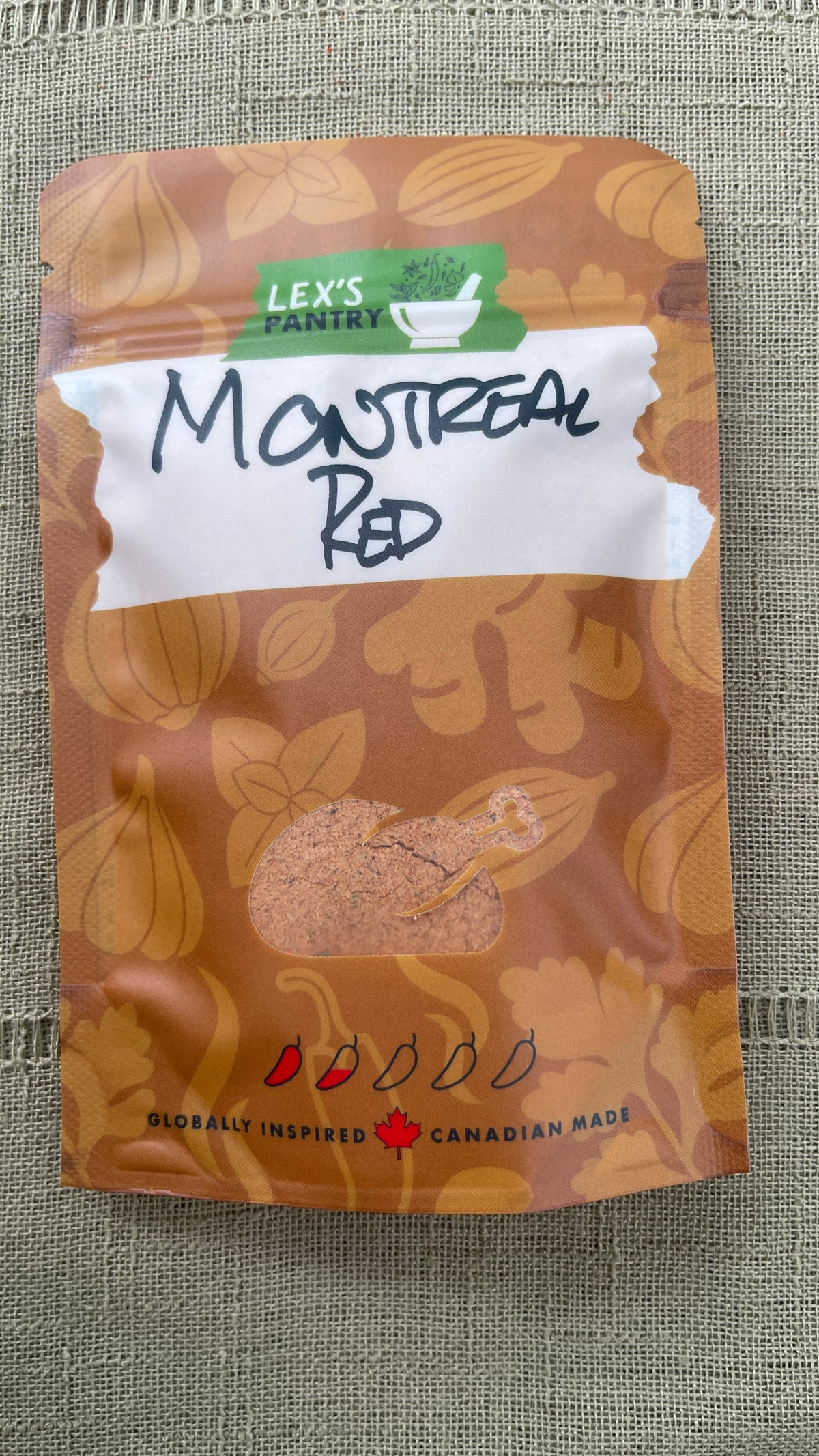 A stand-up pouch of Lex's Pantry Montreal Red Chicken Rub, featuring a kraft brown patterned background with illustrations of garlic, herbs, chili peppers and ginger, a white label with handwritten text "Montreal Red", and a clear window shaped like a roasted chicken showing the seasoning mix inside.