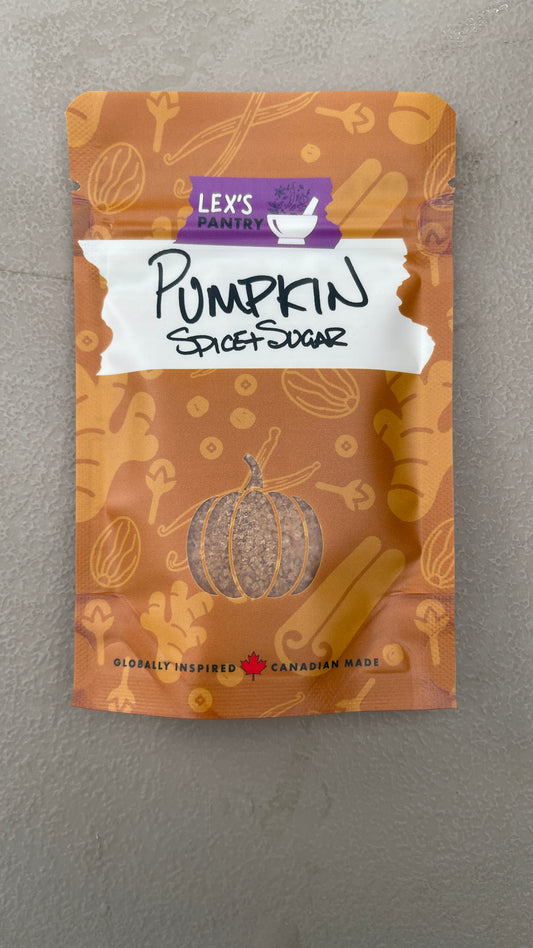 A stand-up pouch of Lex's Pantry Pumpkin Spice+Sugar, featuring kraft-brown design with an assorted spices patterning, a white label with 'PUMPKIN spice+sugar' written in black marker, and a small pumpkin-shaped clear window near the bottom showing the spiced sugar inside.