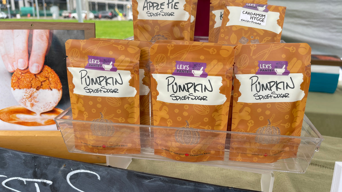 Three bags of Lex's Pantry Pumpkin Spice+Sugar displayed in a clear plastic tray at an outdoor market.
