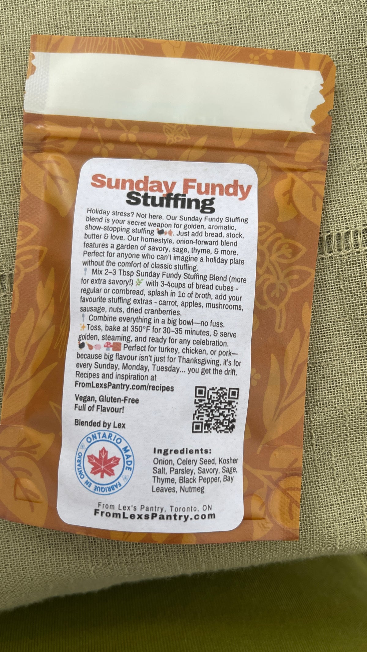 A bag of Lex Pantry's 'Sunday Fundy Stuffing' shown from the back, featuring a large white label with red and black text, a list of ingredients, a QR code and an Ontario Made logo, set against a textured olive green fabric background.