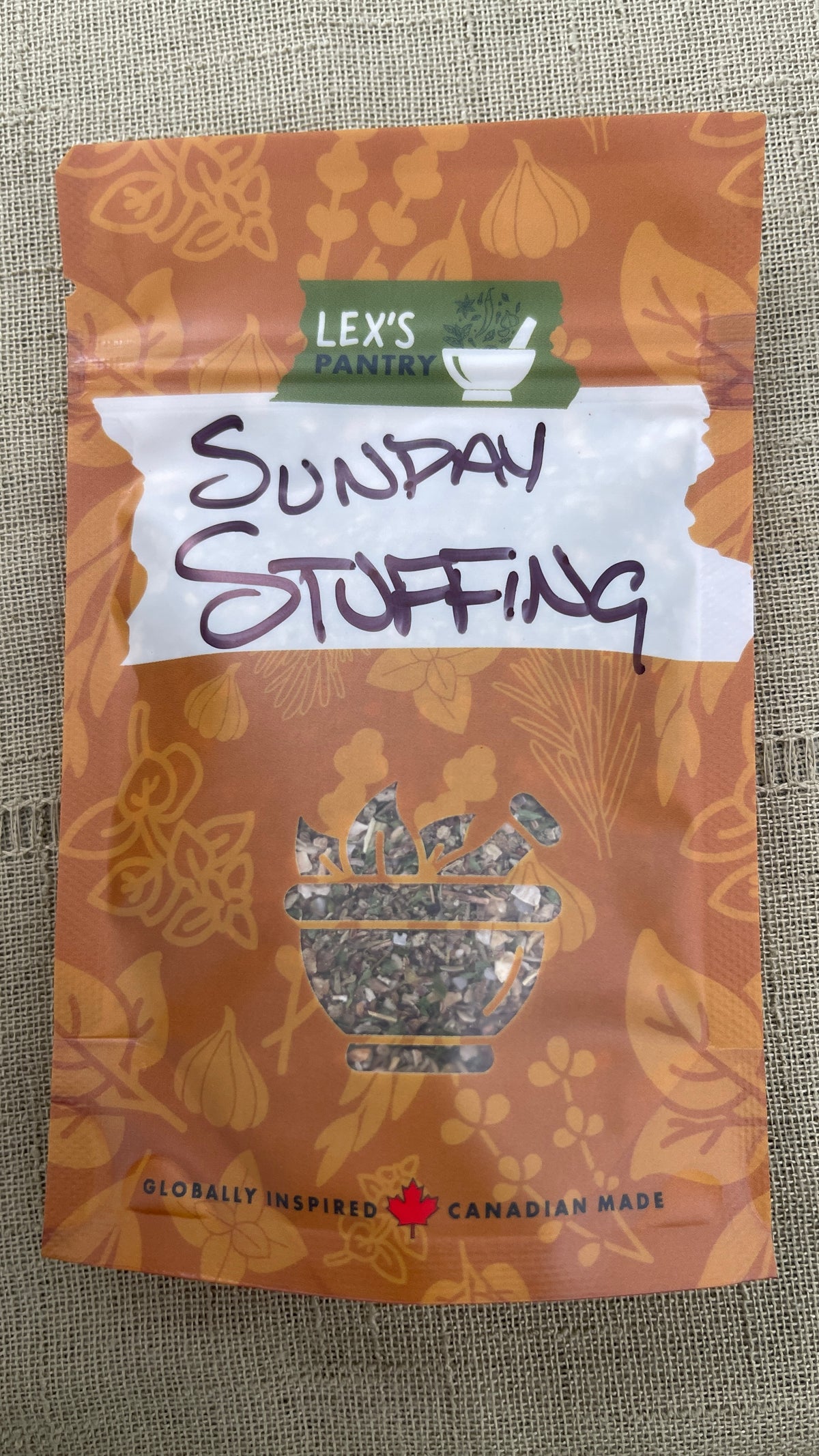 A stand-up pouch of Lex's Pantry's Sunday Fundy Stuffing Seasoning Blend on a textured green fabric background. The packaging is kraft-brown with botanical illustrations of the herbs and garlic inside. It also featuring a white banner with the product name in black handwriting and a green brand label at the top.