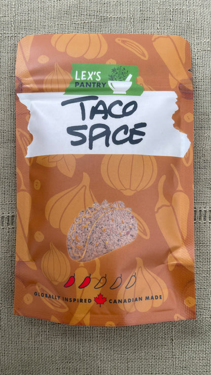 A bag of Lex's Pantry Taco Spice seasoning blend, featuring a brown and orange patterned package with illustrations of onions and peppers, on a textured green burlap background.