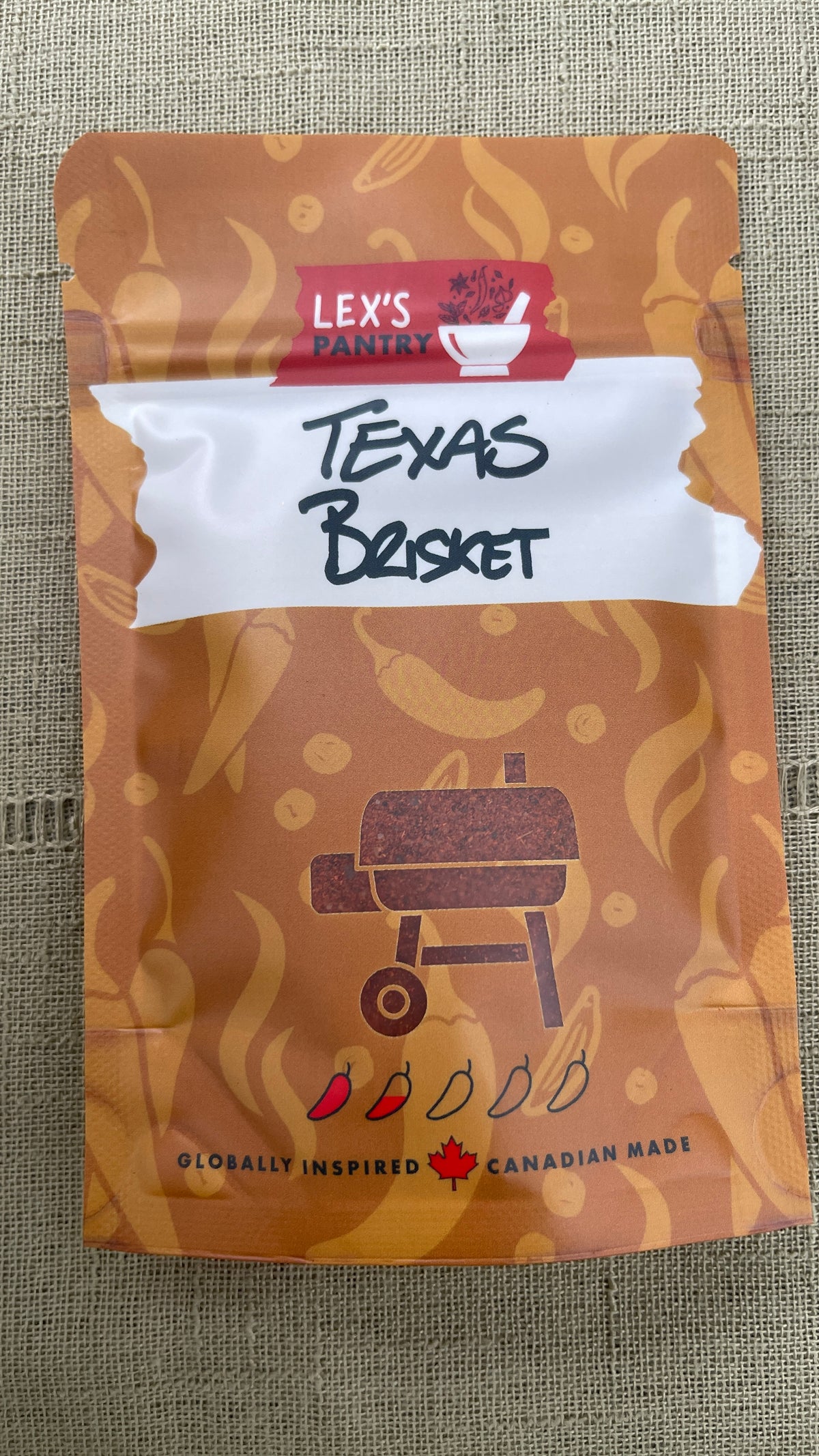 A stand-up pouch of Lex's Pantry Texas Brisket Seasoning and Rub, featuring an Kraft-brown patterned design with a white banner and a barbecue grill clear window showing the spice in the package, placed on a textured green fabric background.