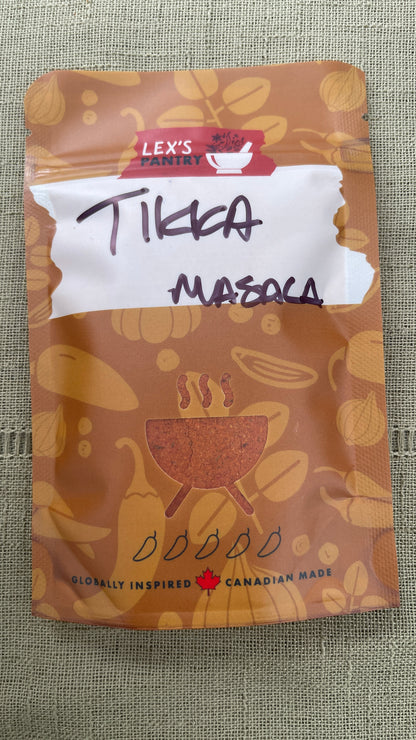 A stand-up pouch of Lex's Pantry Tikka Masala , featuring a brown design with illustrations of spices and a white label area with handwritten text, placed on a textured beige fabric background.